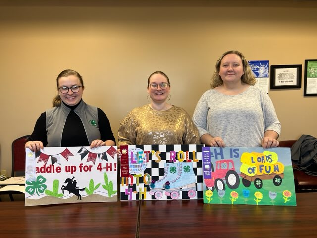 Three women holding 4-H posters. They are titled Saddle up for 4-H, Let's Roll and 4-H is Loads of Fun.