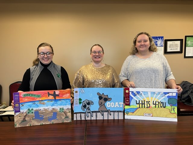 Three women holding posters from a 4-H contest. They are titled Dive into 4-H, 4-H is the Goat and 4-H is 4-You