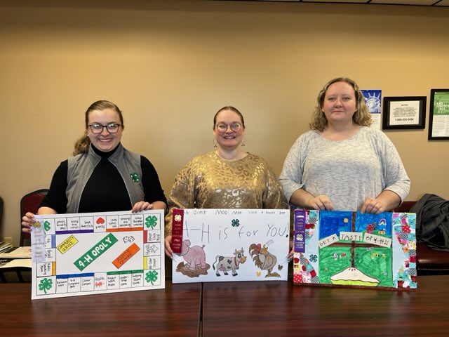 Three women holding 4-H posters. They are titled 4-H opoly, 4-H is for you and Get a taste of 4-H.