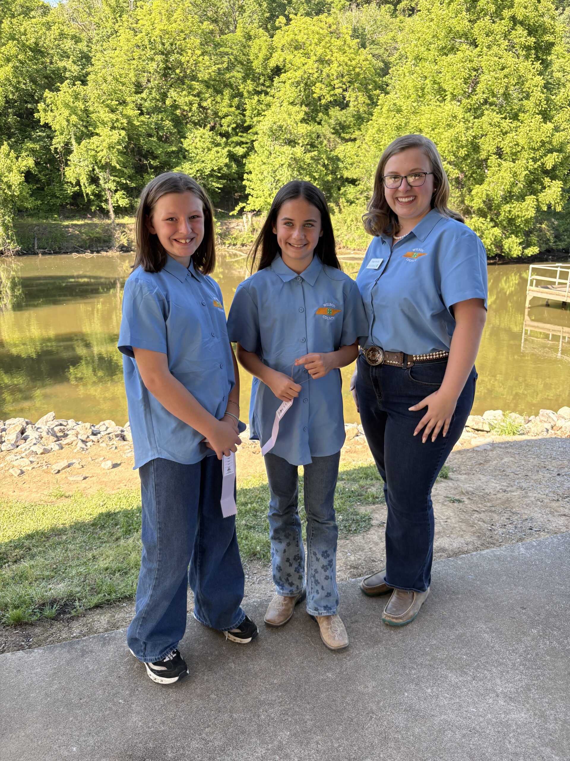Two 4-H youth standing in front of a body of water with a 4-H Agent. Each youth is holding a ribbon from the contest. 